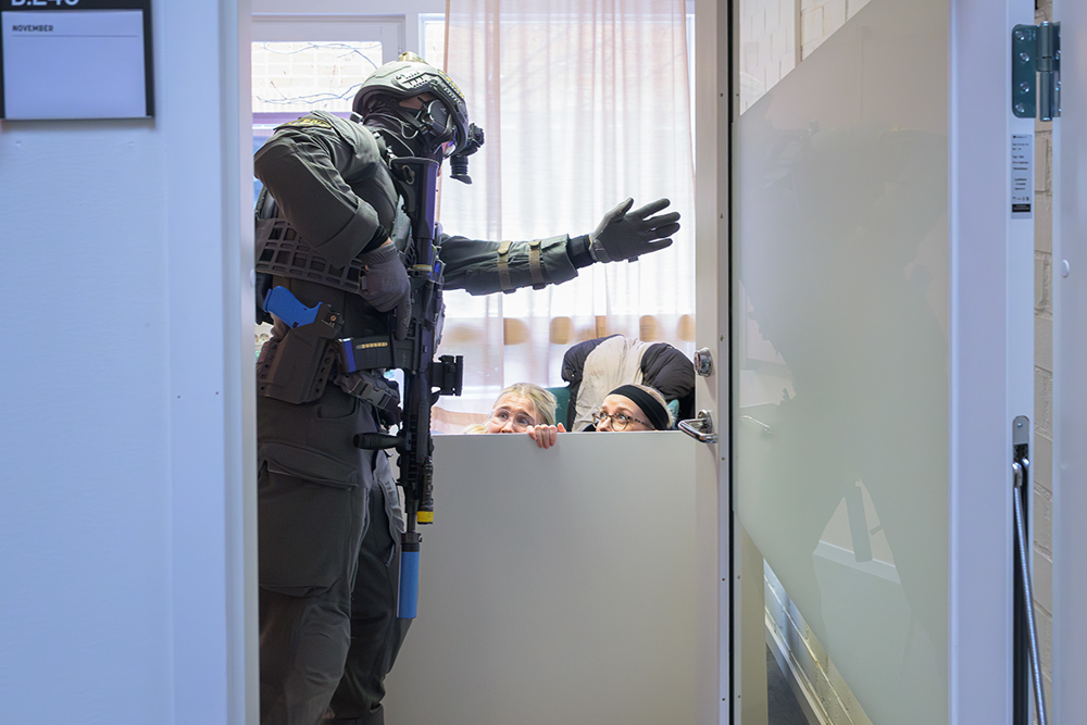 En operatör från insatsstyrkan Öst ber två figuranter att att lämna ett rum där de har tagit skydd bakom ett bord. Foto. tömma rummen på personer som sökt skydd.