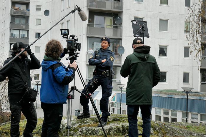 Rissa Seidou står framför ett lägenhetskomplex med ett filmteam framför sig under en filminspelning. Foto.