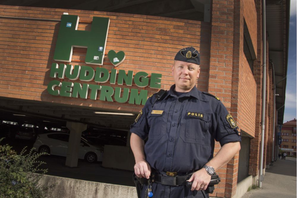 Polisen Reine Berglund står framför en tegelvägg, med en skylt med gröna bokstäver där det står Huddinge centrum