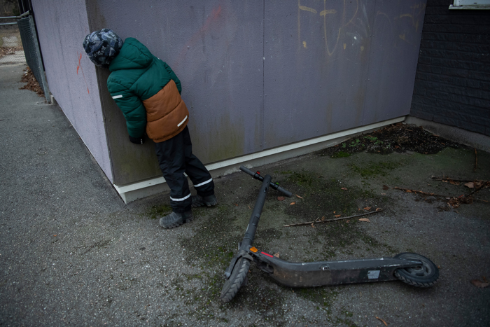 Ett barn i täckkläder och mössa tittar försiktigt fram bakom ett hörn på ett hus.  Arrangerad bild.