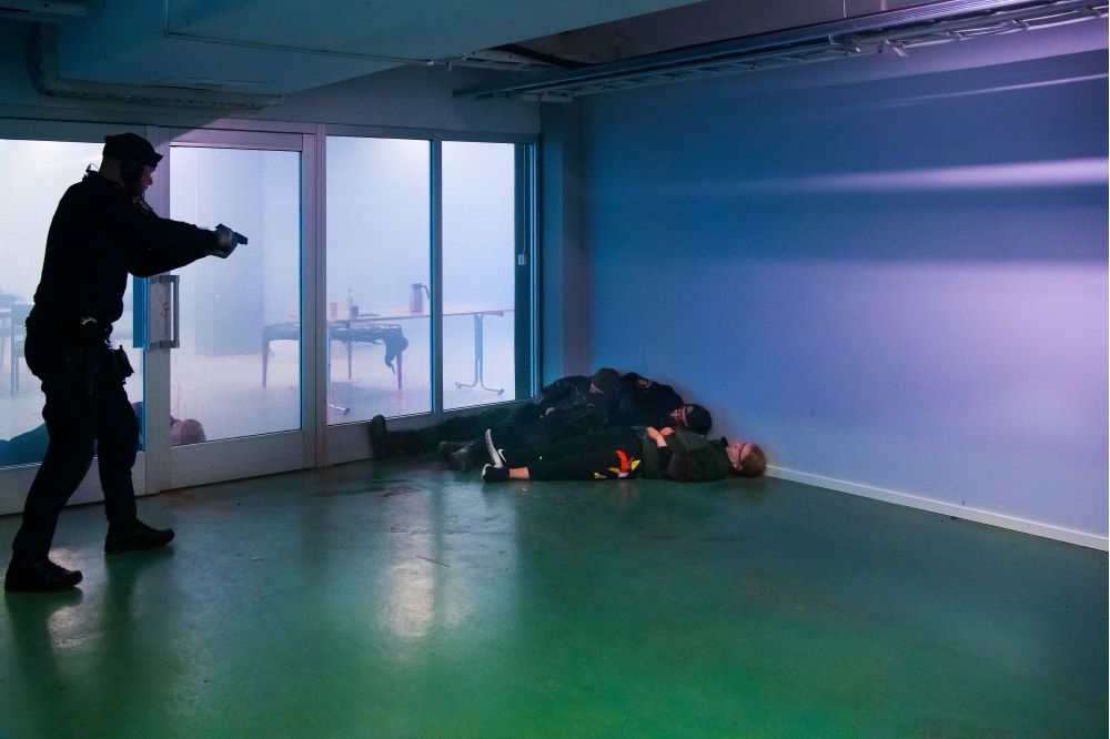 En polis riktar sitt vapen mot en hög av människor på golvet, där gärningspersonen ingår. Foto.