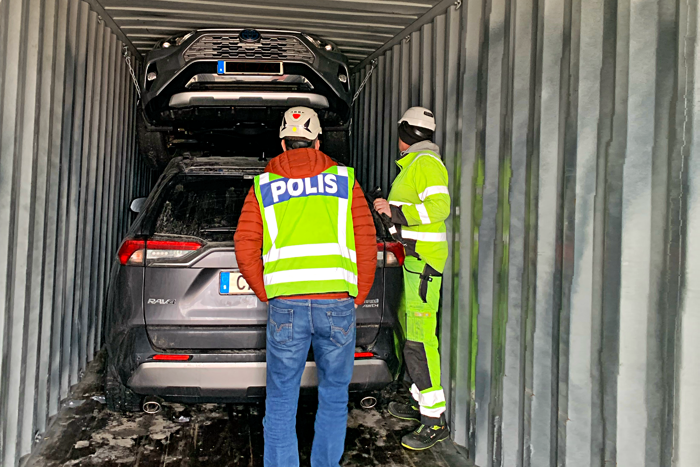 En civilklädd polis i gul väst står och tittar på stulna bilar i en container. Foto.