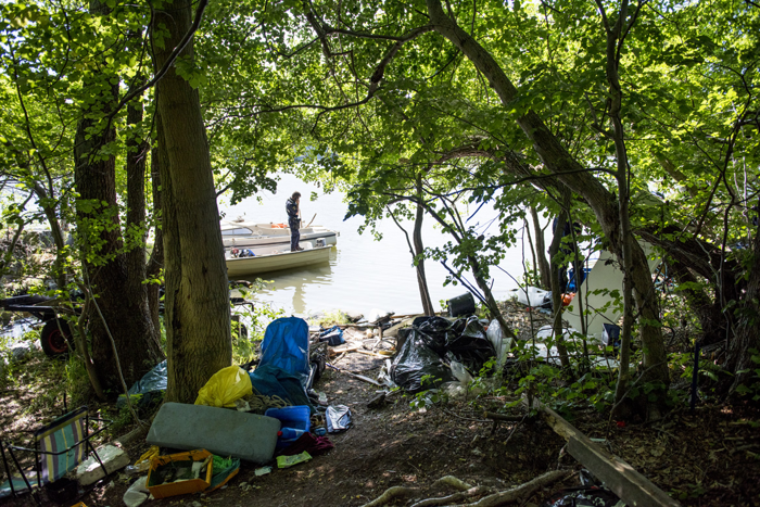 Genom en inramning av träd med sopor längs med marken skymtas sjöpolisen Samuel Sjöö stå i en liten motorbåt. Foto.