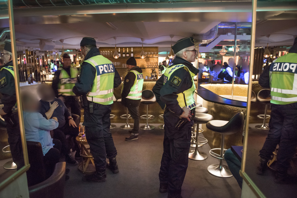 Fyra poliser i gula reflexvästar står vid en bardisk. Två av poliserna pratar med en kvinna och en man, med utsuddade ansikten, som sitter på varsin stol.