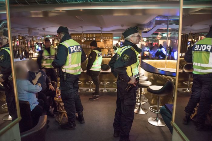 Fyra poliser i gula reflexvästar står vid en bardisk. Två av poliserna pratar med en kvinna och en man, med utsuddade ansikten, som sitter på varsin stol.