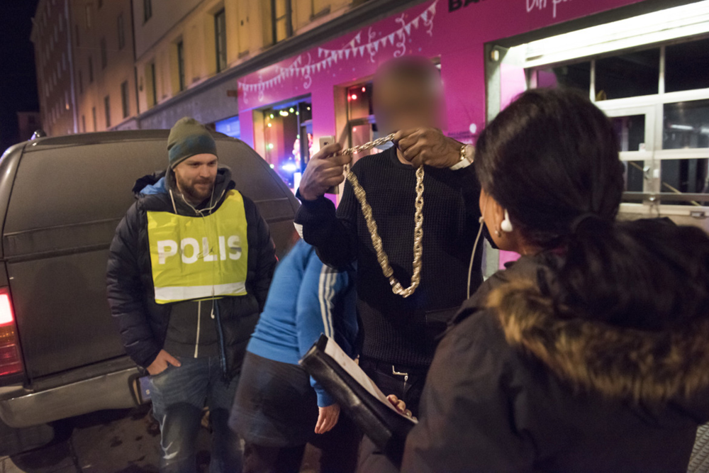 En person, med utsuddat ansikte, står utanför krogen och håller upp en stor guldkedja framför två civilklädda poliser.