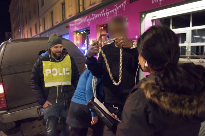 En person, med utsuddat ansikte, står utanför krogen och håller upp en stor guldkedja framför två civilklädda poliser.